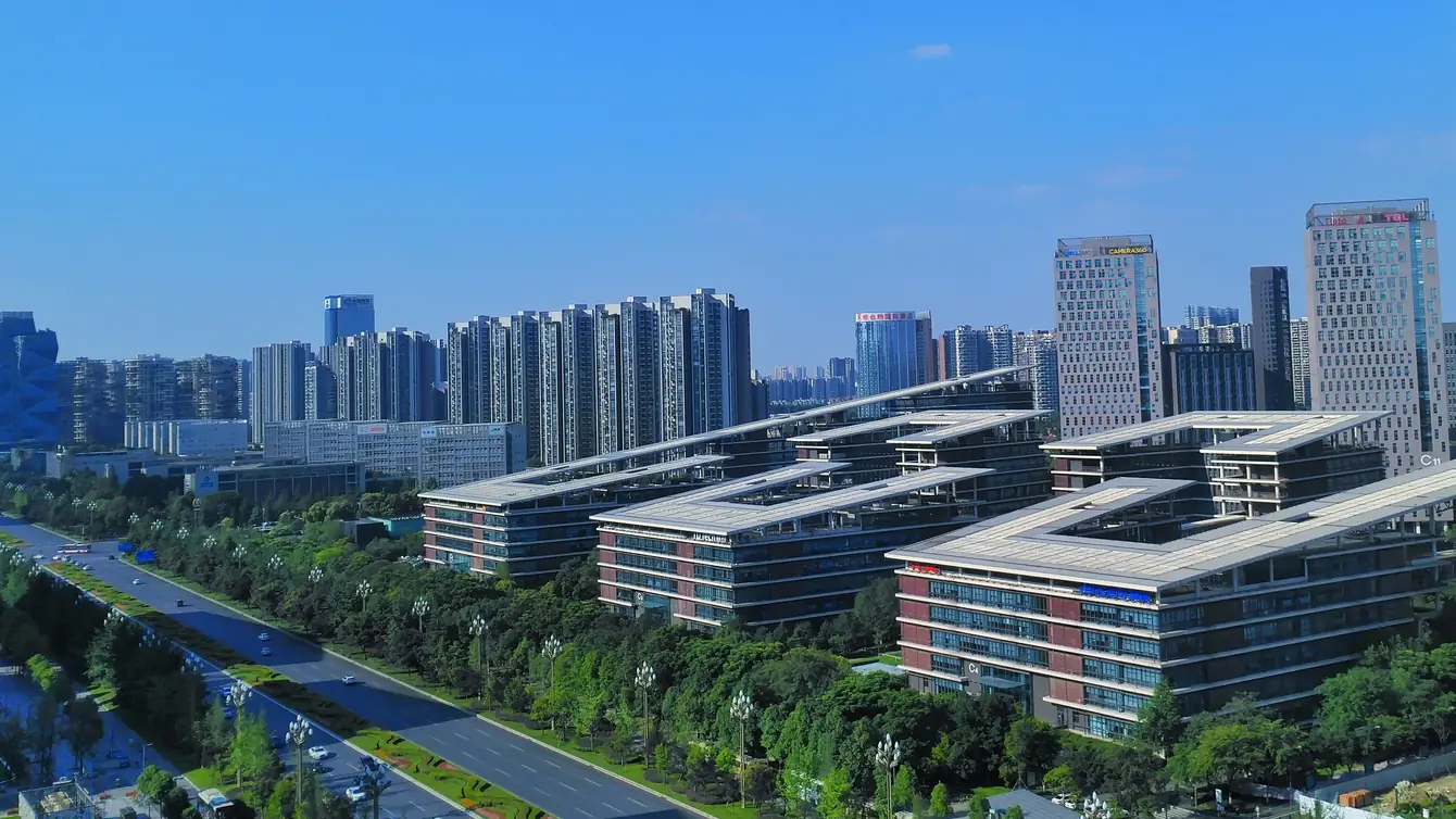 An elevated view of Chengdu’s Tianfu Software Park: long office blocks with stepped, angular roofs set among trees, a wide boulevard running along the left and residential towers beyond, under a clear blue sky