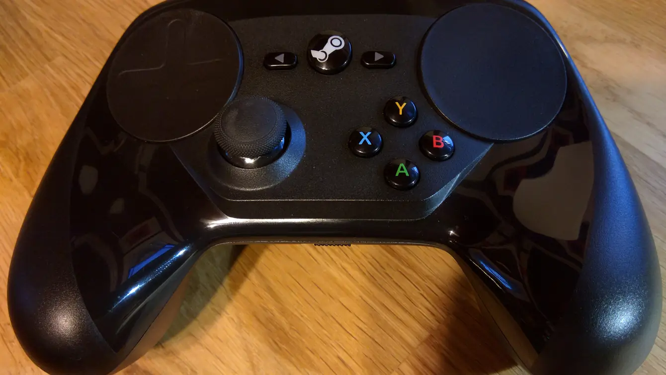 Valve’s Steam Controller seen from above on a wooden table: a black gamepad with two large circular trackpads, a thumbstick, a central Steam button and coloured A, B, X and Y buttons
