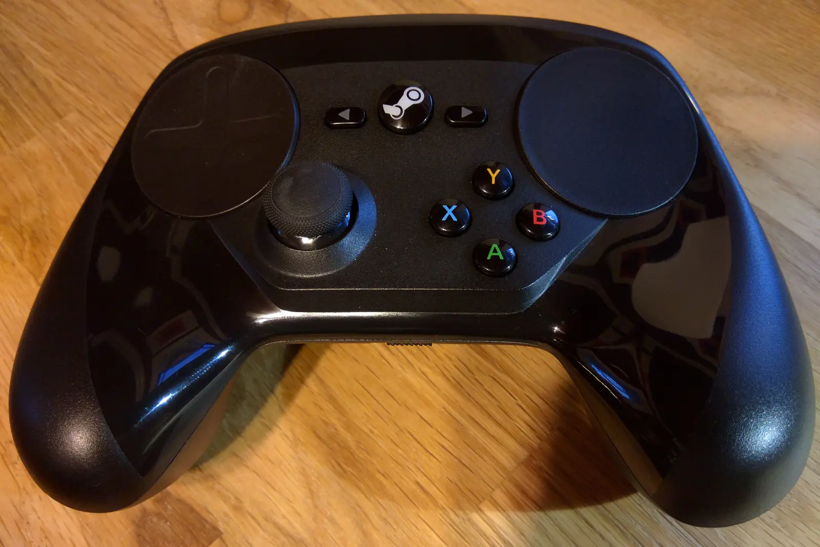 Valve’s Steam Controller seen from above on a wooden table: a black gamepad with two large circular trackpads, a thumbstick, a central Steam button and coloured A, B, X and Y buttons